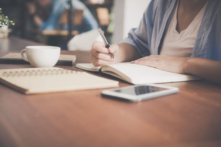 Person writing in a notebook beside a coffee cup and phone